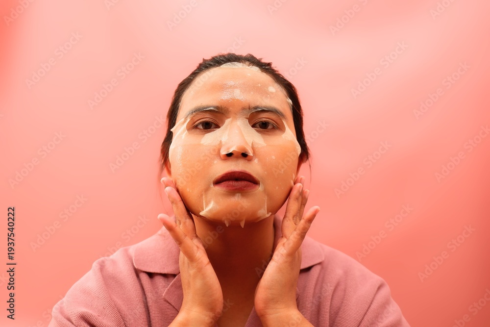 Fototapeta premium Beautiful Asian woman applying a sheet mask for facial hydration and skincare treatment.