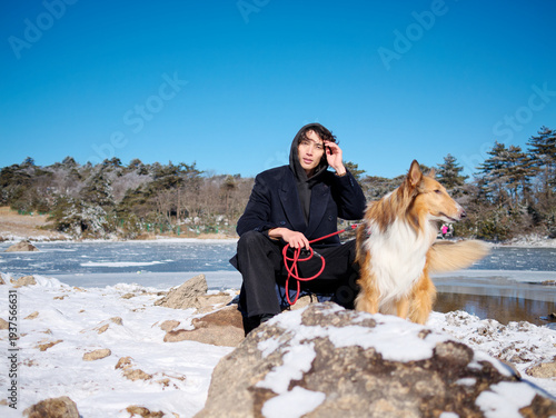 Handsome Chinese young man with curly hair in black overcoat sitting with his dog on snow covered frozen lake in sunny winter day, looks like old friends. Adventure and travel lifestyle with pet.