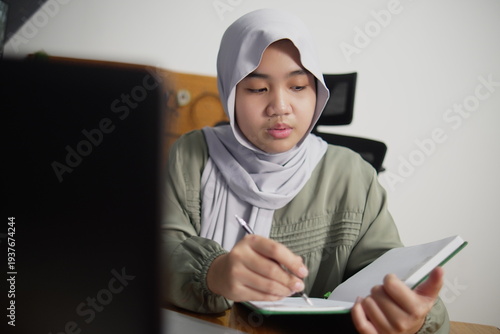 Happy Asian Muslim teenage girl studying with laptop and writing on notebook in her bedroom