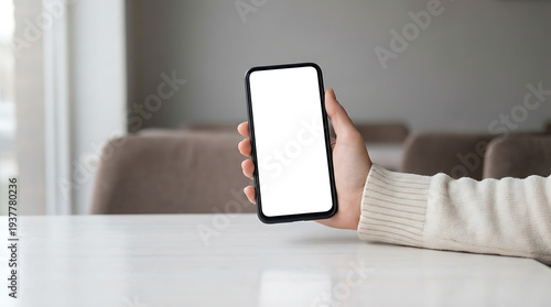 Hand holds phone with blank white screen vertically, resting on white table indoors