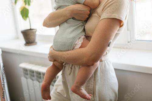 Side view of a woman in light homewear with a Continuous Glucose Monitoring on her arm holding a baby while standing at home near a window.