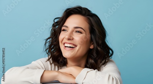 Joyful woman with dark wavy hair laughs heartily against a solid pastel blue backdrop.