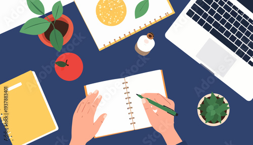 Flat lay of a desk with a notebook, laptop, plants, and a hand writing, creating a productive and creative workspace