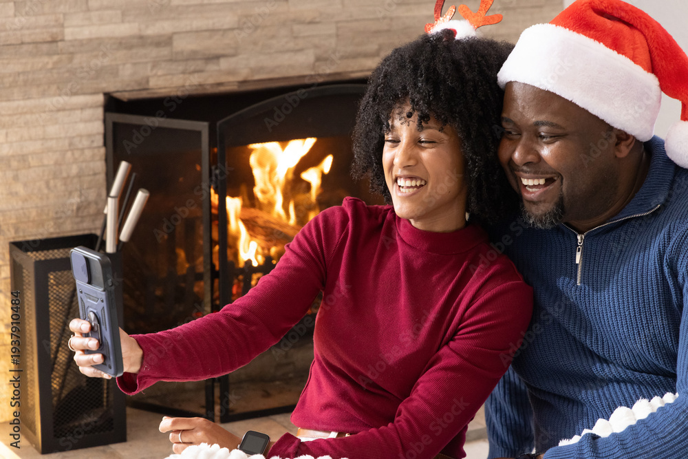 Obraz premium African American couple taking selfie in living room by lit fireplace with smartphone and Santa hat