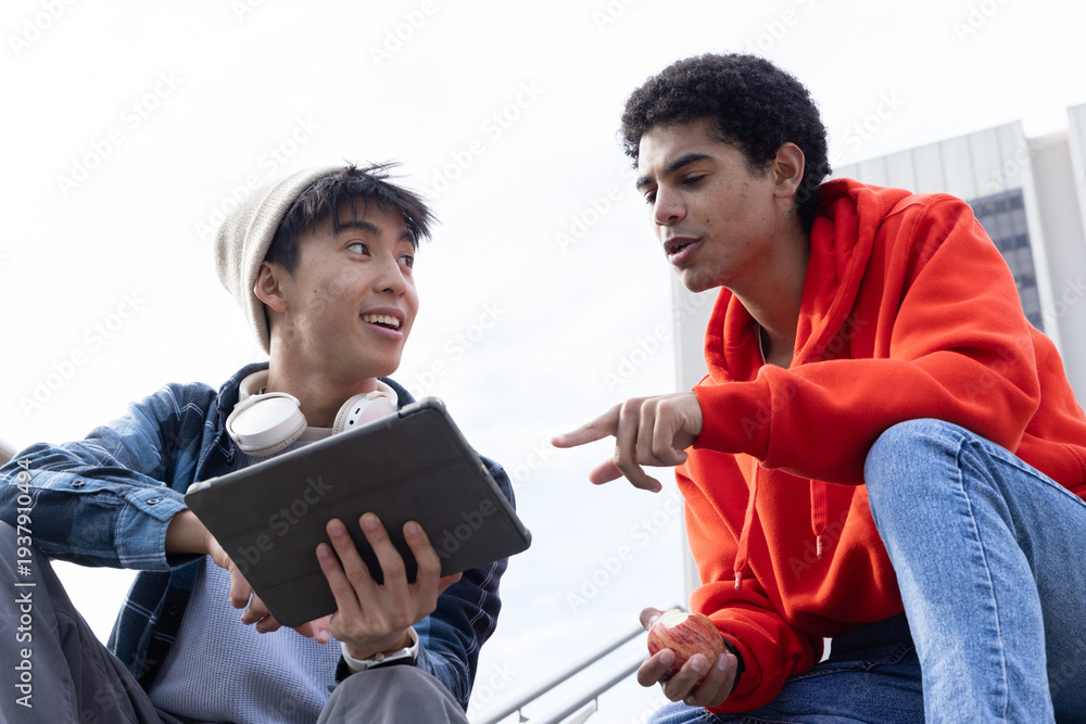 Fototapeta premium Diverse male teenagers sitting on campus steps, pointing at tablet, holding apple and headphones