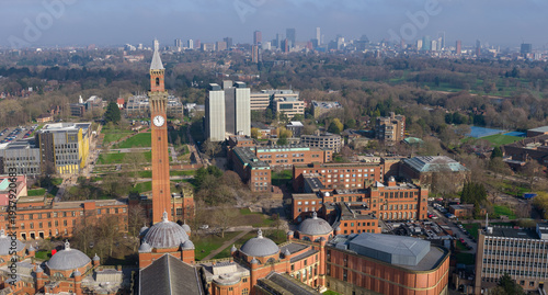 Aerial panorama featuring the Joseph Chamberlain Memorial Clock Tower on the University of Birmingham campus, with a hazy metropolitan skyline and lush greenery in the distance.