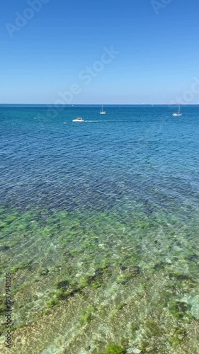 Clear blue sea with small boats on the horizon and transparent shallow water near the shore on a sunny day.