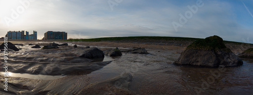 Tableau sur toile Low tide exposes wet sand, rocks and shallow tide pools along a coastal shoreline with a grassy sea wall and distant waterfront apartments beneath a calm, cloudy dusk sky