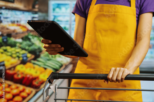 Grocery store worker in yellow apron holds tablet over shopping cart in produce aisle, checking inventory or online order fulfillment, with fresh vegetables in background.