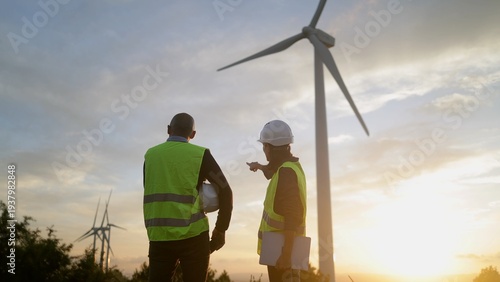 Quadro su tela Engineers in safety vests overlooking a wind turbine farm at sunset, discussing