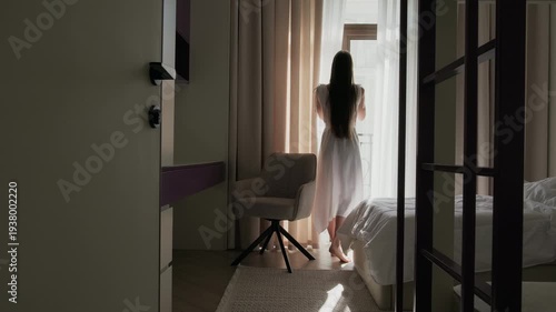Woman closing curtain and walking away from window in minimalist bedroom interior. Modern apartment with loft bed above sofa, soft natural light and neutral color palette. 