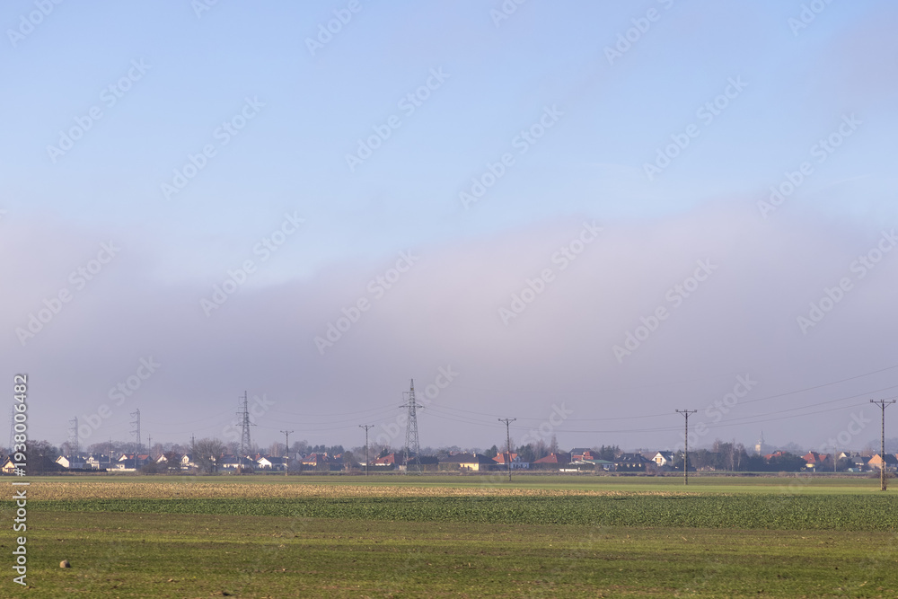 Fototapeta premium Village houses and prominent electricity pylons emerge from low-lying mist across green fields, highlighting rural energy infrastructure under a clear sky