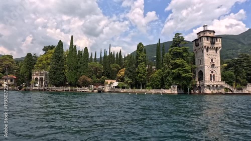 Torre San Marco at Gardone Riviera on Lake Garda from a boat, stone tower beside cypress gardens above rippled water with green hills under summer clouds