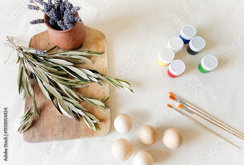 Easter still life in a rustic style: American brown chicken eggs, fresh lavender in a ceramic pot and olive branches on a wooden cutting board, brushes and colored paints on a white flax background.