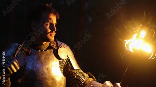 A knight in full armor, his face bloodied and scarred, holding a sword and torch, is sneaking through a dark night forest. A medieval warrior searches in the darkness.