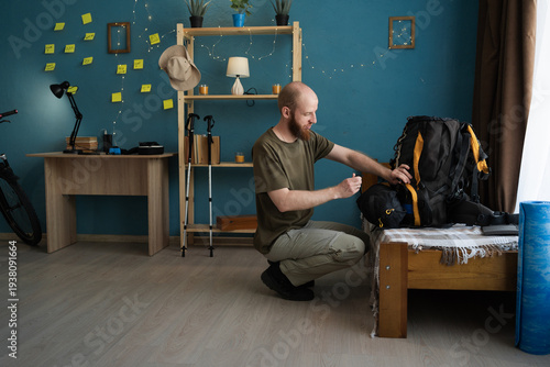 Bearded young man packing trekking backpack for hiking journey or expedition in home living room. adventure, tourism, travel, hiking and active lifestyle.