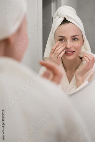Bilde på lerret Portrait of young woman with towel on head applying under eye patches making dermatology procedure near mirror in bathroom