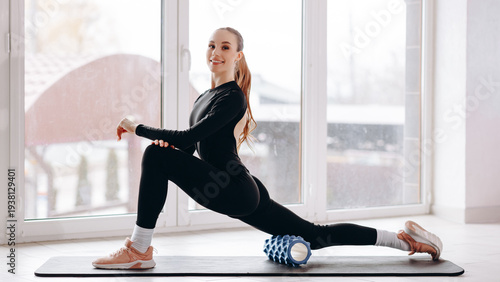 Young woman doing a lunge stretch with foam roller during an indoor fitness workout by window