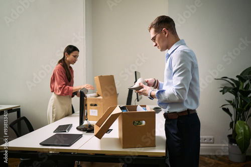 Young Caucasian man and young Asian woman cleaning office desks by packing items into cardboard boxes, both focused on organizing workspace, modern office environment visible