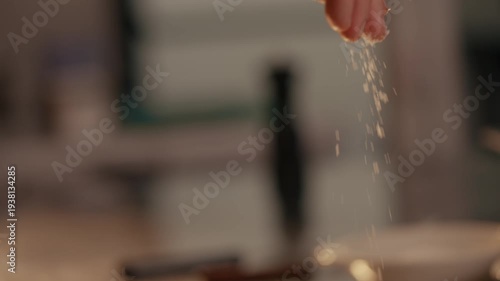 Hand sprinkles coarse salt over raw steak. Close-up captures salt falling. Kitchen background softly blurred. Action focuses on seasoning meat. Ideal for cooking, food, or restaurant content