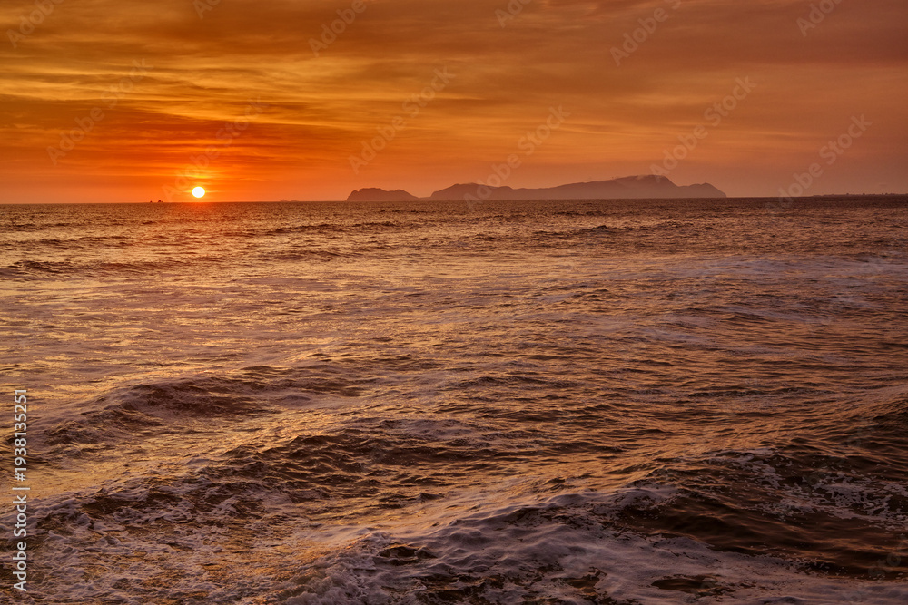 Obraz premium A vibrant summer sunset over the Pacific Ocean along Lima’s Costa Verde, with the silhouette of San Lorenzo Island on the horizon. Warm golden light reflects across the waves