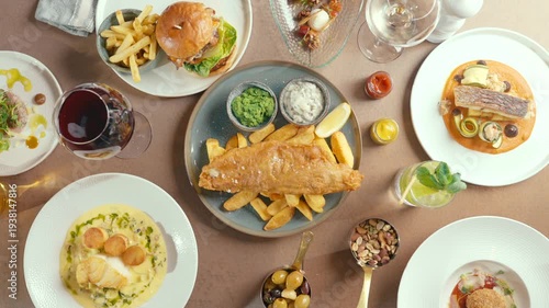 Overhead view of people serving fish and chips on a rustic table. Hands reach for food with casual confidence. Diverse dishes with colorful sides and drinks surround the central plate