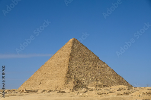Large stone pyramid rising above an open desert landscape, its geometric shape standing sharply against the clear blue sky with sunlight highlighting the sandy terrain around its base