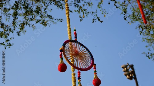Circular handmade hanging ornament with colorful threads, beads, and red tassel pots gently swaying in the wind against a clear blue sky and tree branches.