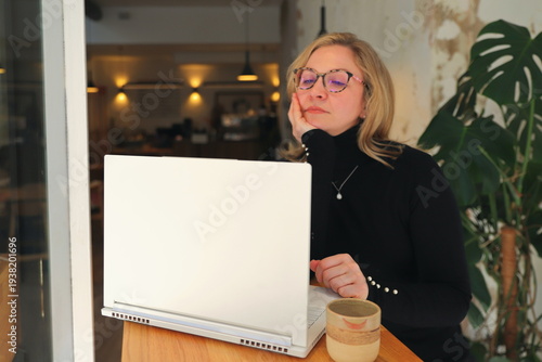 Woman wearing glasses and a black turtleneck looks at her phone while working on a laptop