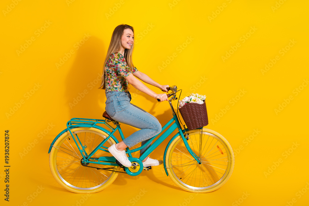 Fototapeta premium Young woman rides a bright turquoise bike with flowers in a basket against a bold yellow background