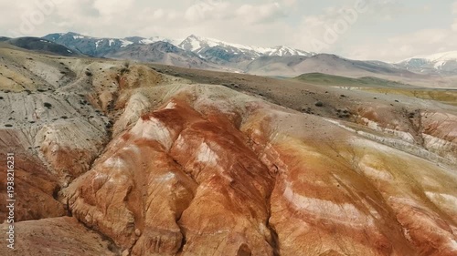 A colorful landscape featuring layered rock formations in shades of red, orange, and gray. Snow-capped mountains rise in the background under a cloudy sky.