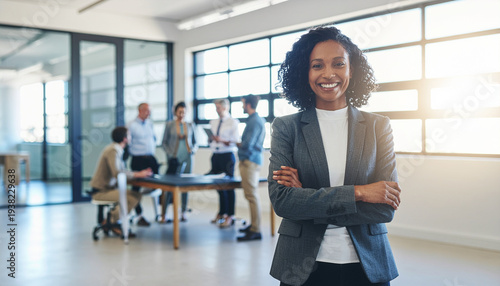 Confident Black businesswoman smiling with arms crossed in a modern office, colleagues working in the background.