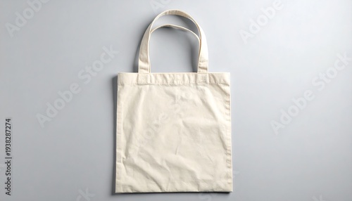 Beige fabric tote bag with handles lies flat against a light gray background, casting faint shadows