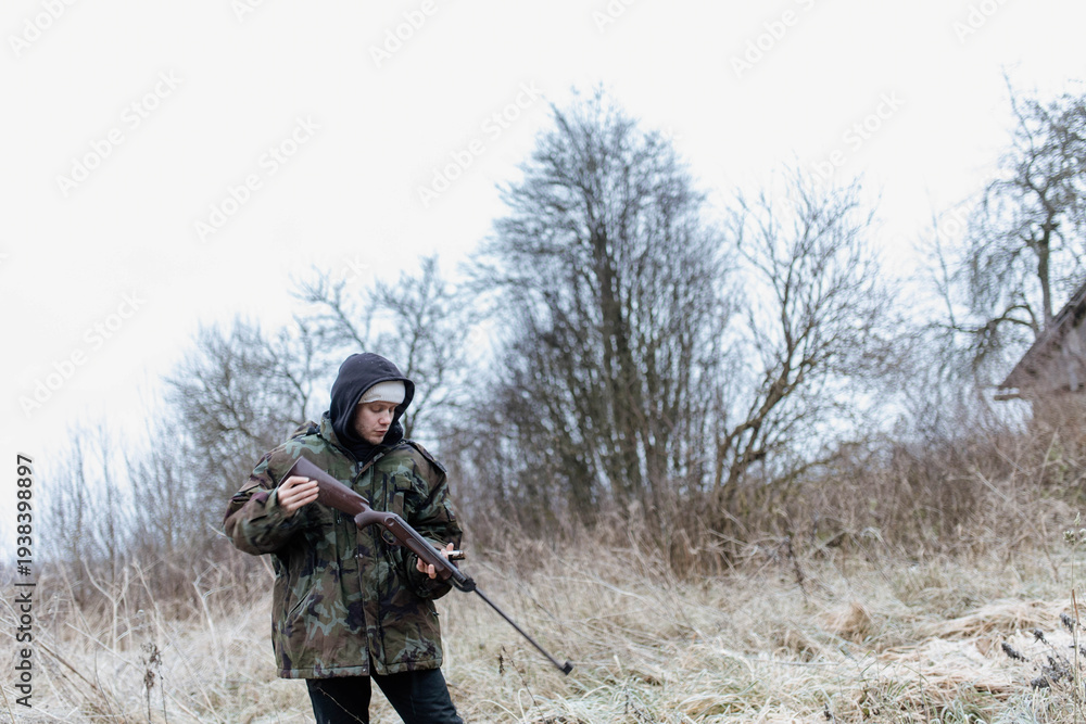Fototapeta premium A male hunter with a gun in a camouflage jacket in a field in cold weather