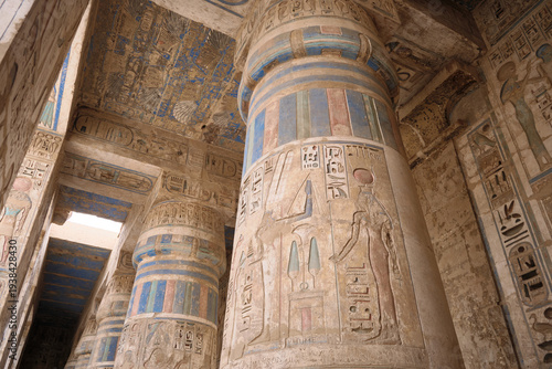 Low-angle view of massive stone columns inside ancient Egyptian temple decorated with carved hieroglyphs and painted reliefs. Medinet Habu, Theban Hills, West Bank of the River Nile, Luxor, Egypt