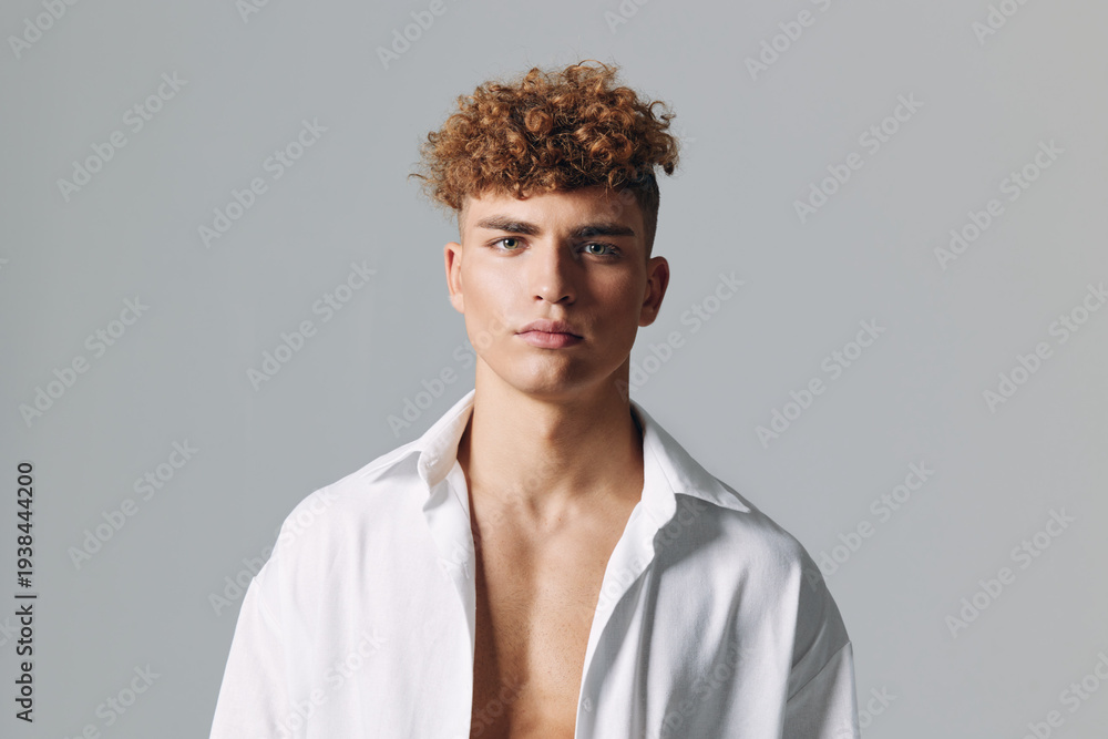 Obraz premium Young man with curly hair wearing open white shirt posing against gray background, looking confidently at camera with neutral expression and relaxed posture.