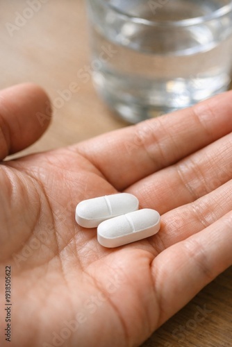Two white pharmaceutical pills resting in an open human hand. A glass of water is visible in the blurred background. Use for healthcare, medical treatment, or wellness content.