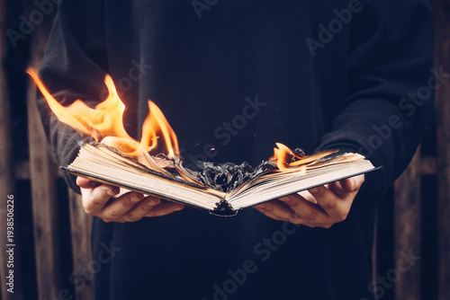 A person dressed in dark clothing holds an open book in flames, its pages curling and turning to ashes. A visual metaphor of destruction and censorship.