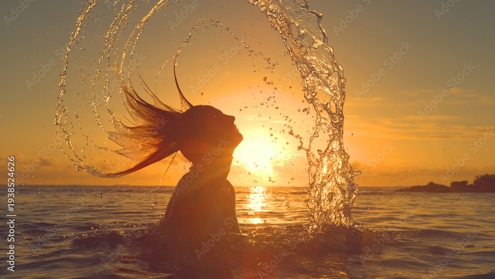 Fototapeta premium SILHOUETTE: Playful woman splashing ocean water by whipping her head backwards.