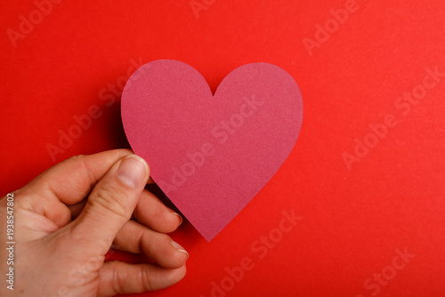 Close up of a hand holding a red heart cutout against a vibrant red background. Perfect for Valentine's Day or love conc