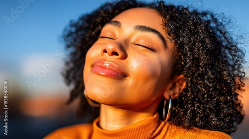 A young Black woman with curly hair smiles with her eyes closed, feeling the warm glow of the golden hour sun on her face. She wears an orange turtleneck and stands outdoors enjoying the moment