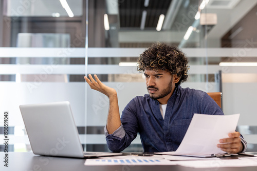Problems with documents and data. A stressed young Indian man sits at a desk in an office and looks at a laptop screen