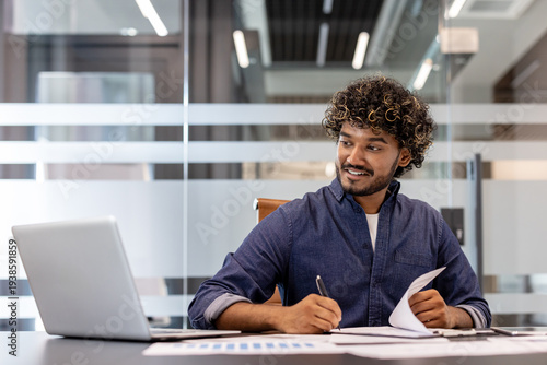 Smiling young Indian man working in the office with documents, looking at the laptop screen and making notes on paper
