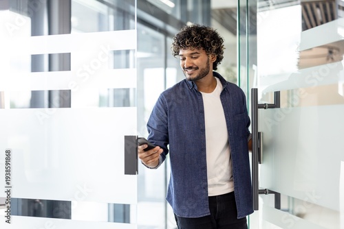 An Indian smiling man holds a phone in his hands, opens the door and enters the office premises