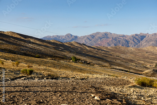 A wide view of the dry Bolivian highlands, showing sunlit brown hills leading to craggy mountain ranges under a clear sky.