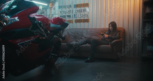Young Asian Woman in Leather Gear Relaxing on Sofa, Scrolling Through Photos on Smartphone After Exciting Ride Near Sport bike. Concept of Motorcycle Hobby and Digital Lifestyle in Garage Workshop.