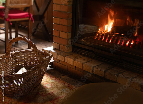 A handcrafted wicker basket used as a firewood holder adds a touch of rustic charm to the setting, next to a rustic brick fireplace with a lit fire.