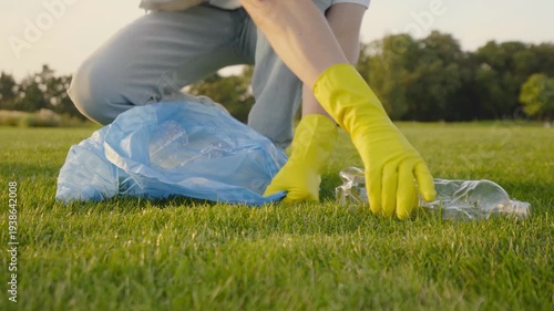 Volunteer in yellow gloves collecting a discarded plastic bottle on green grass in a park, promoting litter cleanup, recycling and environmental care during a community cleanup effort Close-up 4K UHD