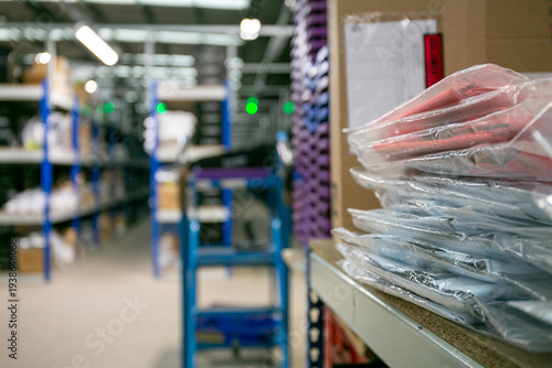 Warehouse storage area with blue metal shelving filled with inventory, packaged items in plastic bags on a shelf, and a blue trolley in the foreground, typical of a fulfilment centre or stockroom