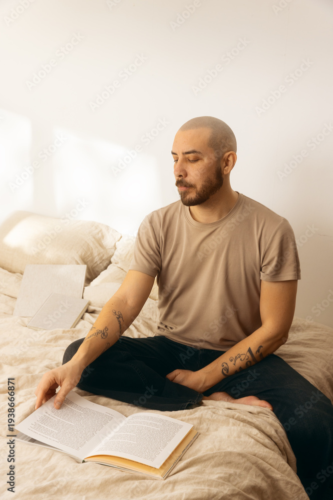 Fototapeta premium Tattooed man with a beard and shaved head sitting cross-legged on a bed, reading an open book in a bright, minimalist room with natural sunlight. Concept of relaxation, peace, and mindful literacy.